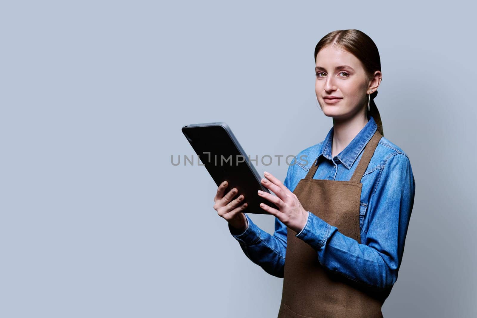 Young smiling female worker in apron uniform holding digital tablet on gray studio background. Digital technologies in business work, Internet apps applications for online orders and customer service