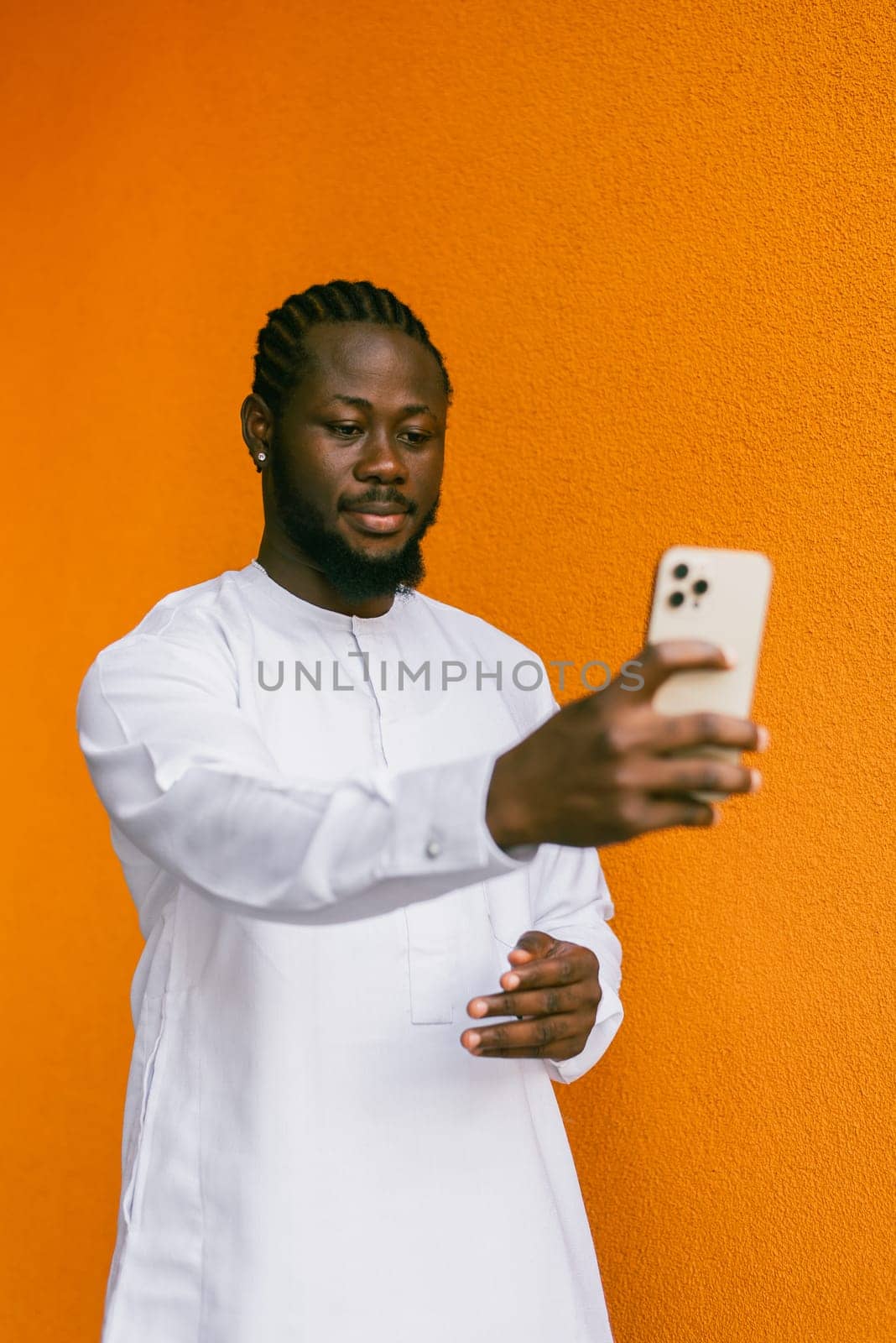 Happy young African American man in dashiki ethnic clothes taking selfie on orange wall background. Millennial generation student and youth. by Satura86