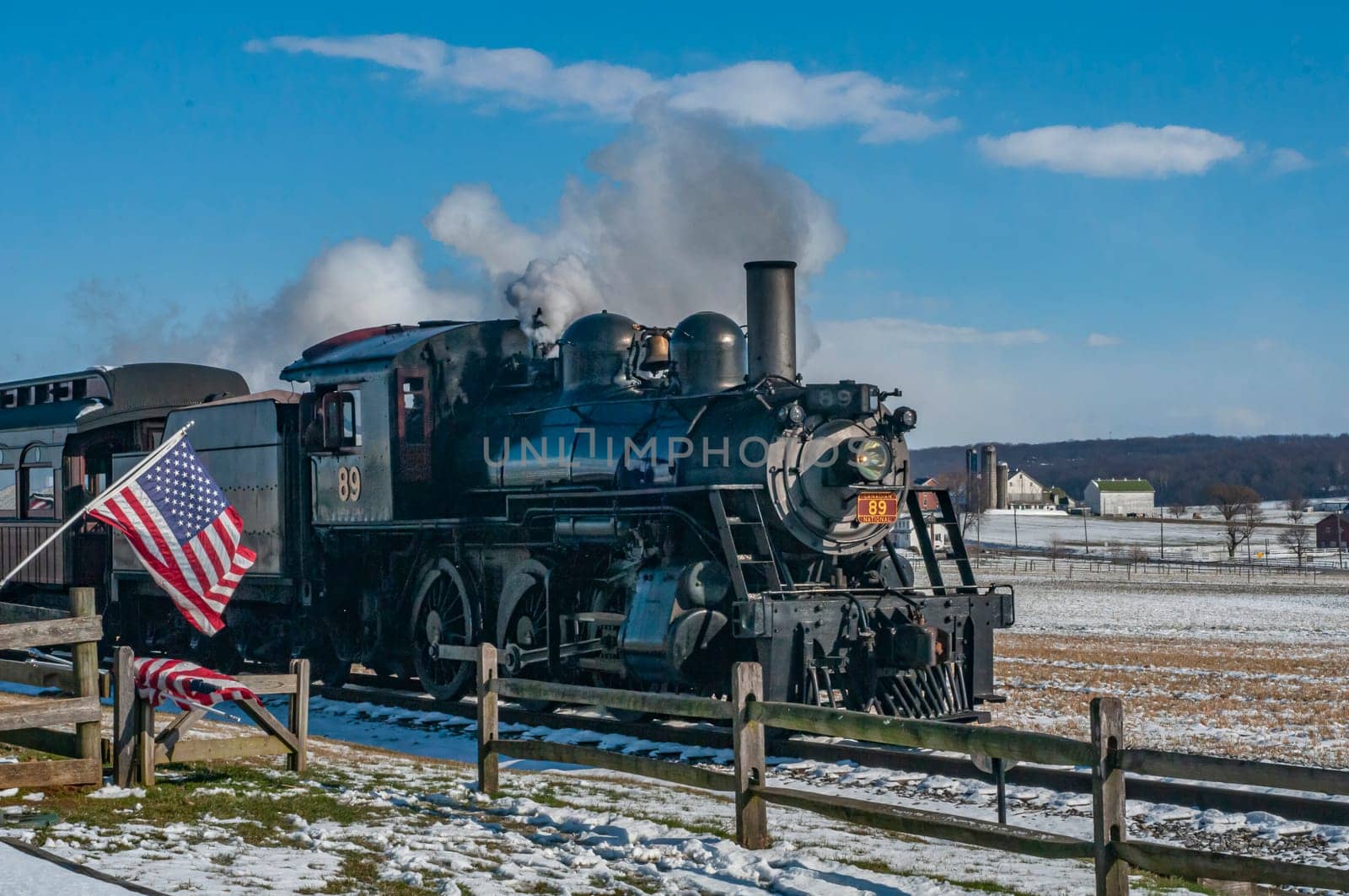 Ronks, Pennsylvania, USA, February 17, 2024 - A steam train is pulling a train car with an American flag on it. The train is traveling through a snowy field