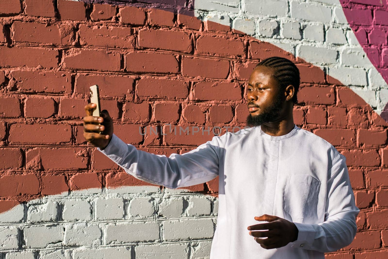 Happy young African American man in dashiki ethnic clothes taking selfie on brick wall background. Millennial generation student and youth. by Satura86