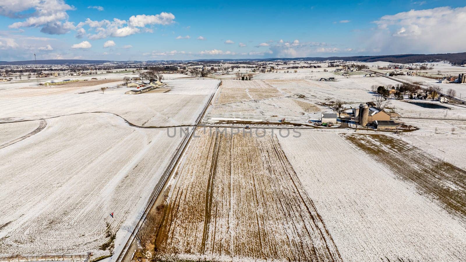 An Aerial View Of Expansive Snow-Dusted Farmland With Rural Buildings And Silos.