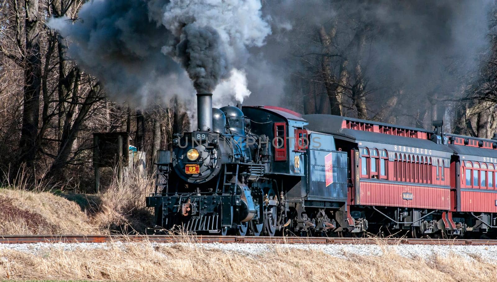 Ronks, Pennsylvania, USA, February 17, 2024 - A steam train is traveling down the tracks, leaving a trail of smoke in its wake. The train is surrounded by trees, and the smoke billows out of the engine, creating a sense of movement and power