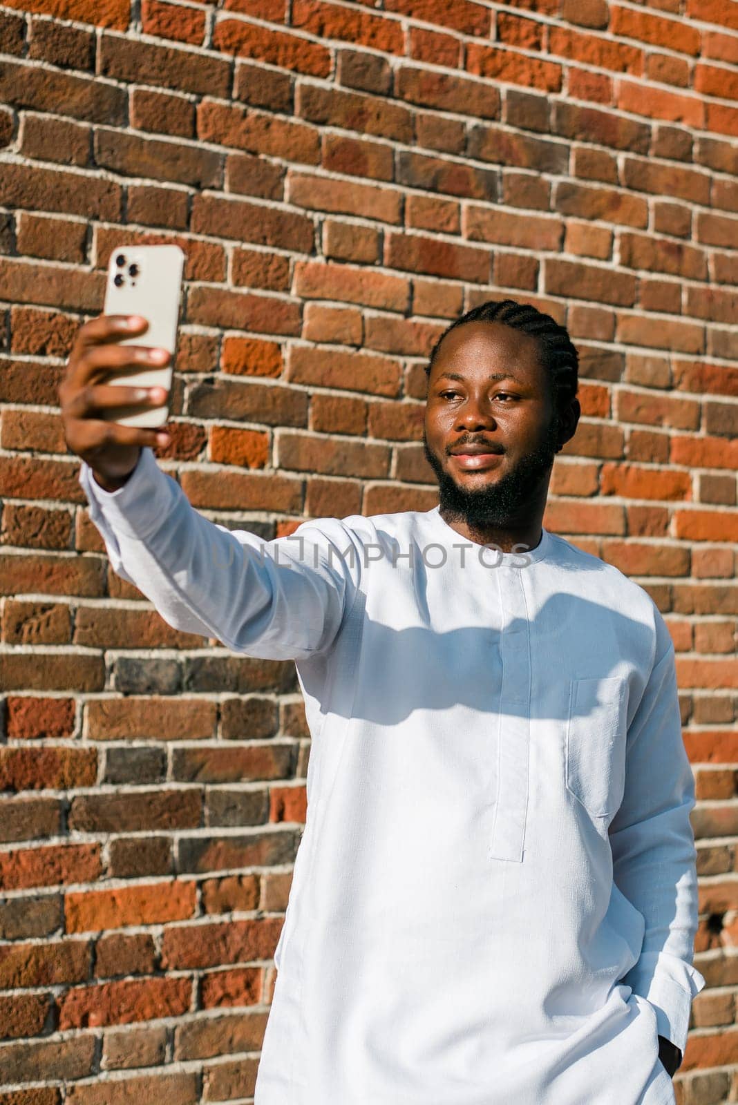 Happy young African American man in dashiki ethnic clothes taking selfie on brick wall background. Millennial generation student and youth. by Satura86