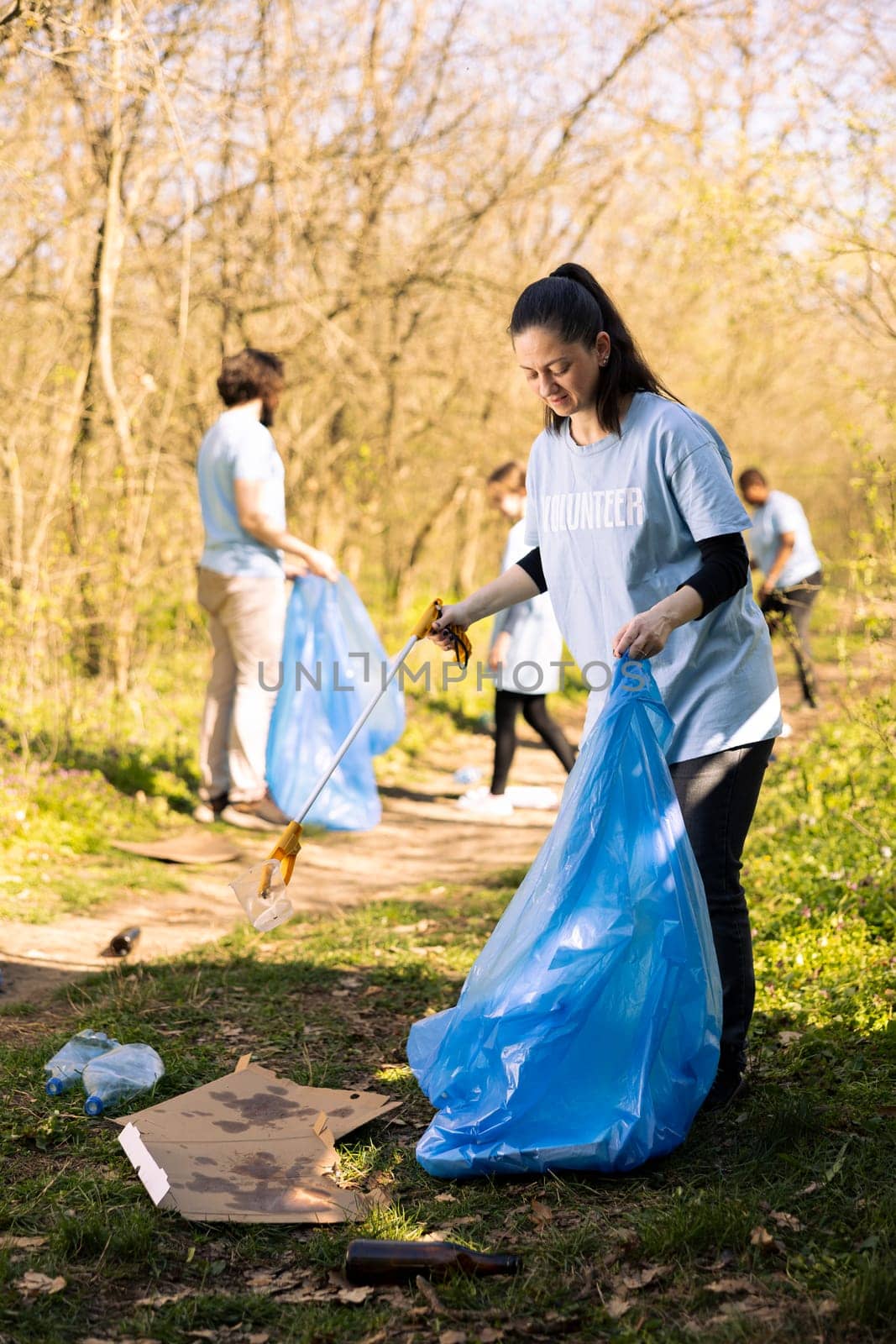 Female volunteer tidying the woodland of garbage and plastic bottles, collecting rubbish with claw and disposal bag. Young activist volunteering to conserve natural ecosystem.