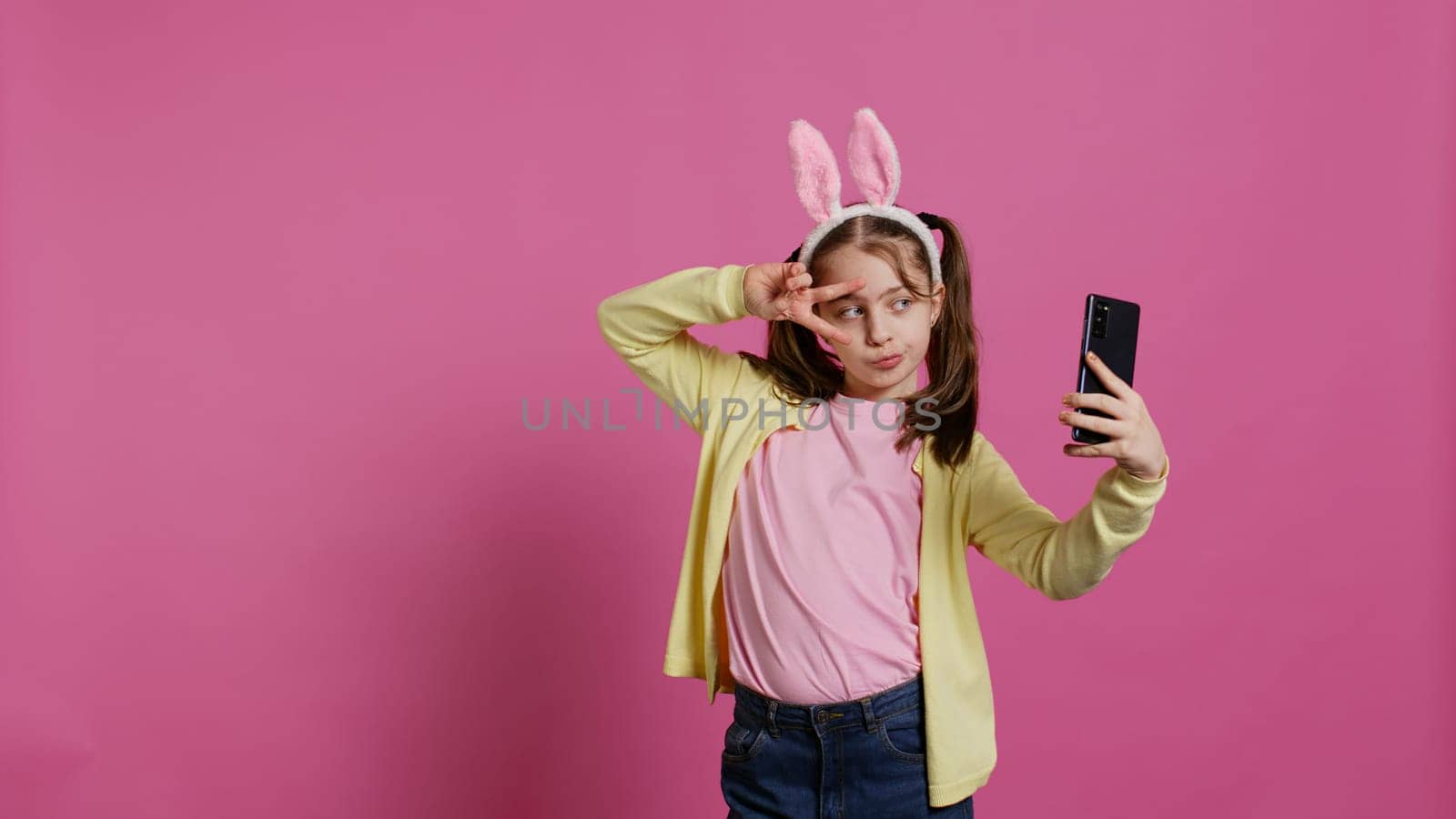 Confident happy young girl taking photos on smartphone webcam, feeling cute with her pigtails and bunny ears. Joyful smiling toddler takes pictures and fooling around in studio. Camera B.