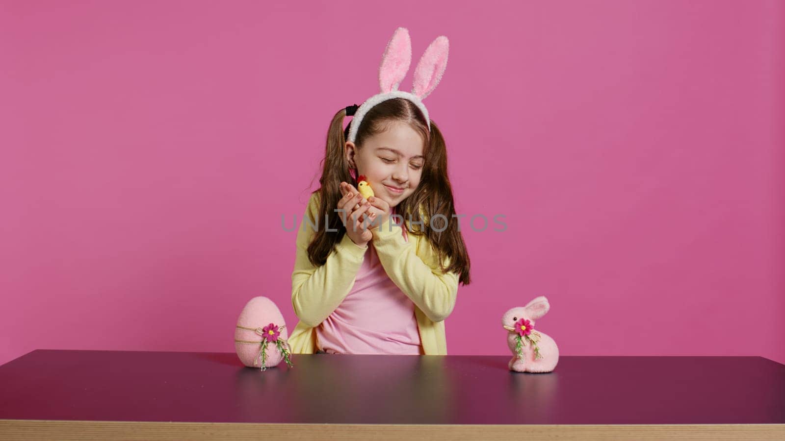 Joyful little girl playing with festive easter decorations in studio, creating arrangements with a chick, rabbit and egg. Smiling cute toddler with bunny ears showing colorful ornaments. Camera B.