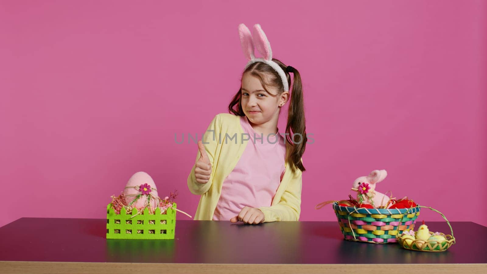 Happy schoolgirl with bunny ears showing thumbs up sign, feeling excited about easter holiday preparation. Cheerful child doing like okay sign and decorating baskets for festivity. Camera B.