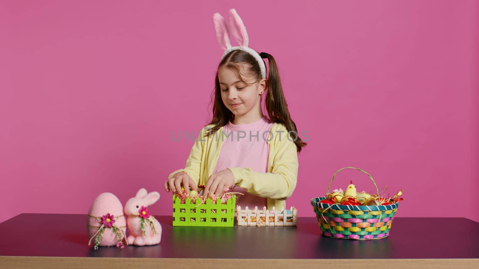 Joyful little girl showing her handcrafted festive basket on camera, creating a colorful handmade arrangement in time for easter holiday. Small toddler with bunny ears decorating eggs. Camera B.