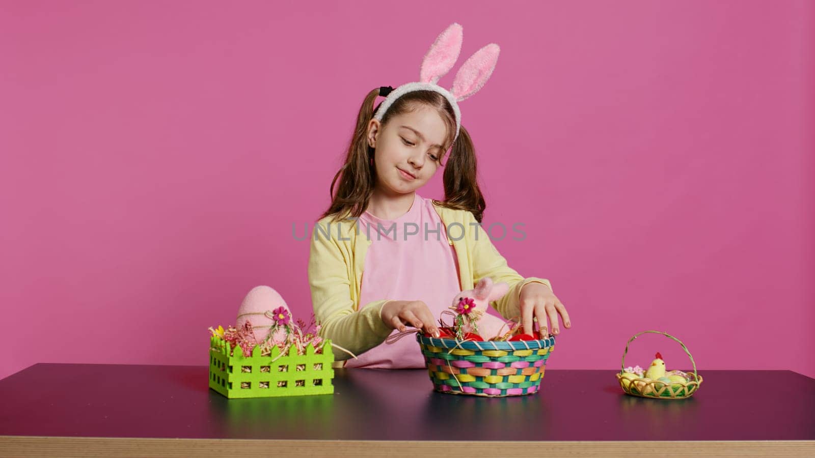 Excited young girl arranging painted eggs in a basket to prepare for easter holiday celebration, creating festive arrangements. Playful happy toddler with bunny ears, creative activity. Camera B.