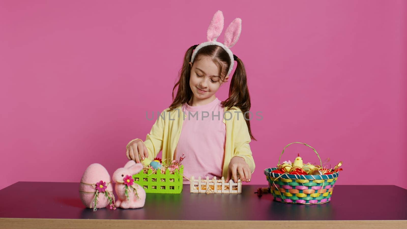 Smiling happy schoolgirl presenting a handmade decorated basket in studio, making easter holiday preparations against pink background. Young child showing handcrafted arrangements. Camera B.
