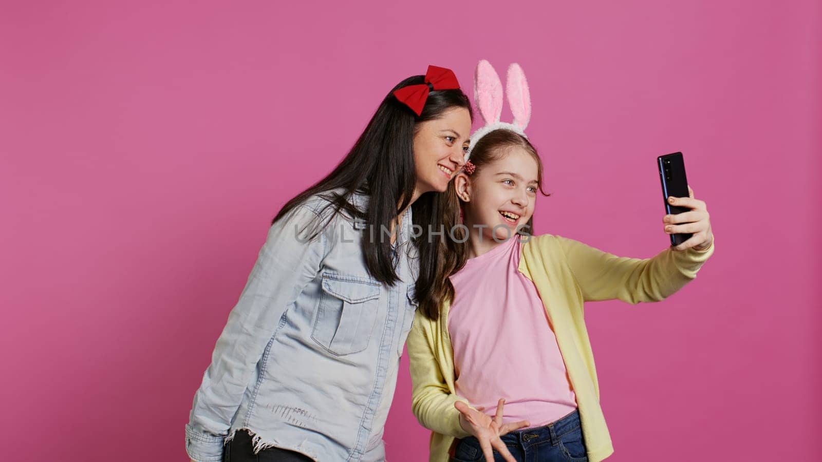 Little schoolgirl and her mom talking on videocall meeting in studio, waving at webcam and chatting with their family. Cute preteen and mother using smartphone for a teleconference chat. Camera B.