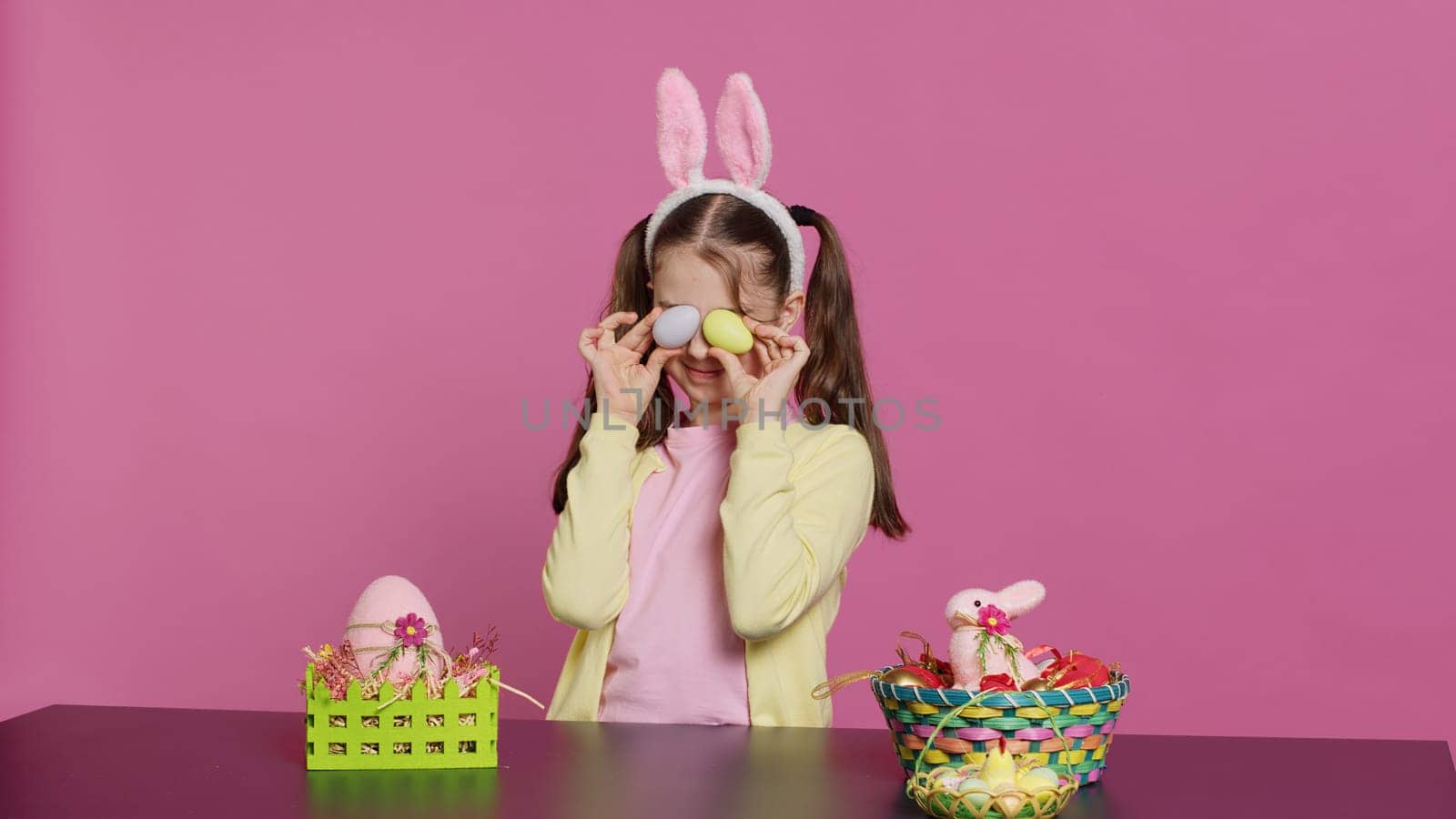 Enthusiastic young girl playing peek a boo in front of camera, using painted easter eggs against pink background. Joyful lovely toddler feeling excited about spring holiday festivity. Camera A.