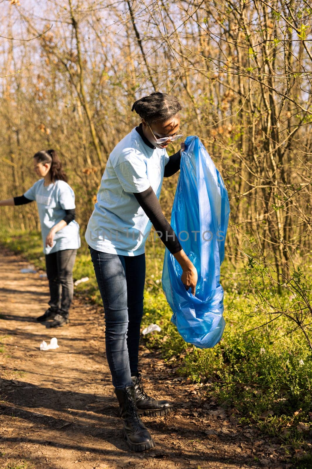 Woman volunteer grabbing junk and plastic waste with a claw tool, clearing natural ecosystem of garbage. African american girl doing voluntary work against illegal dumping.