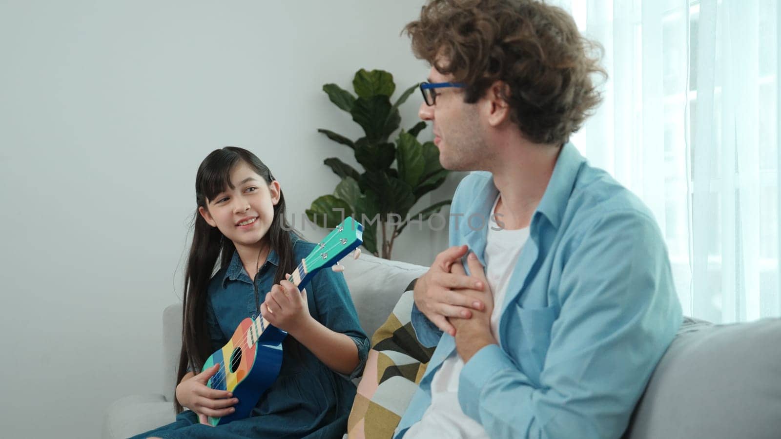 Back view of happy father looking at smart daughter playing ukulele warmly and singing song. Happy dad motivated and encouraged american school girl enjoy playing guitar. Family recreation. Pedagogy.