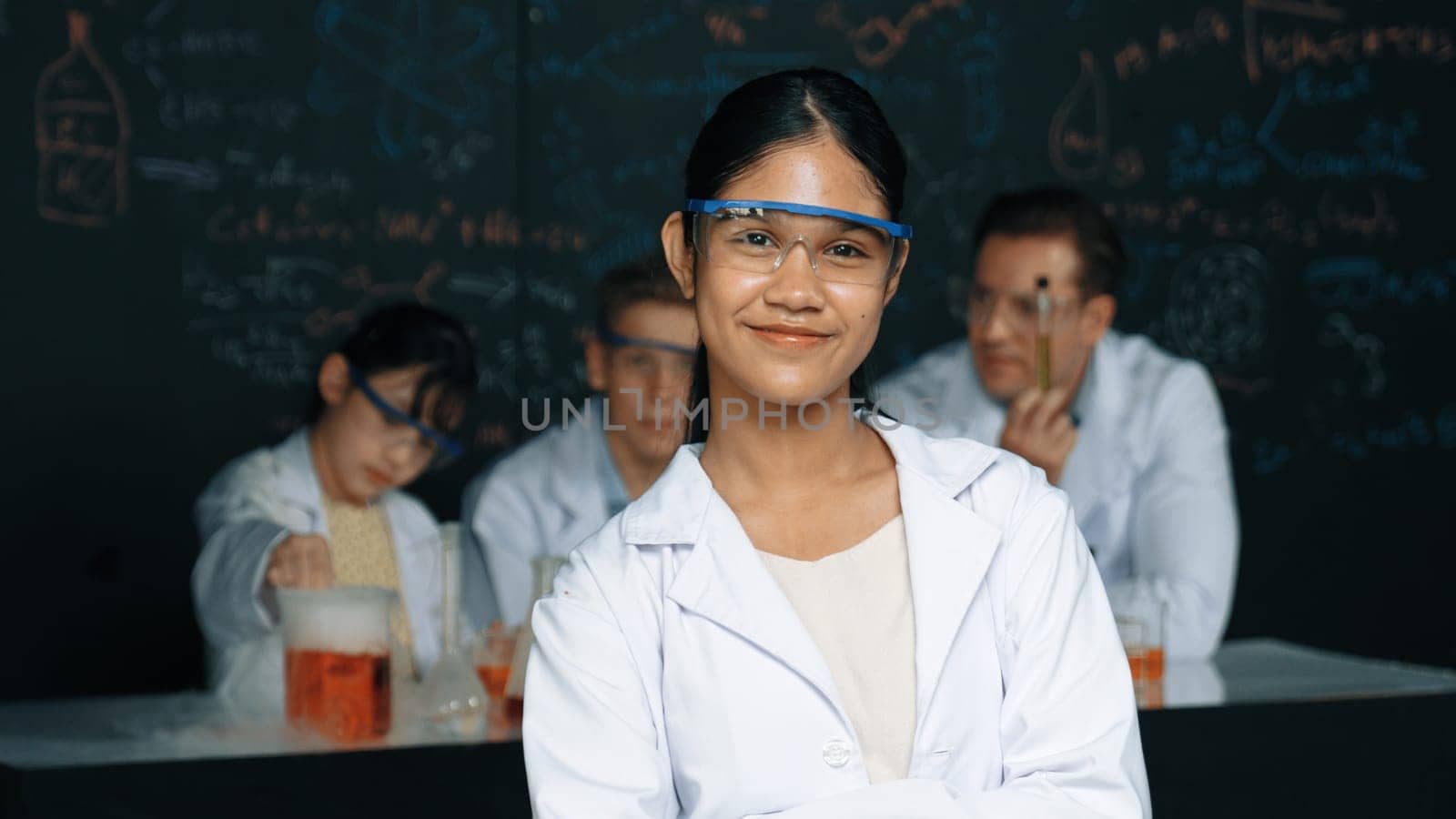 Girl looking at camera with arm folded while people doing experiment at laboratory. Cute student standing blackboard with chemical theory with blurring background at STEM science class. Edification.