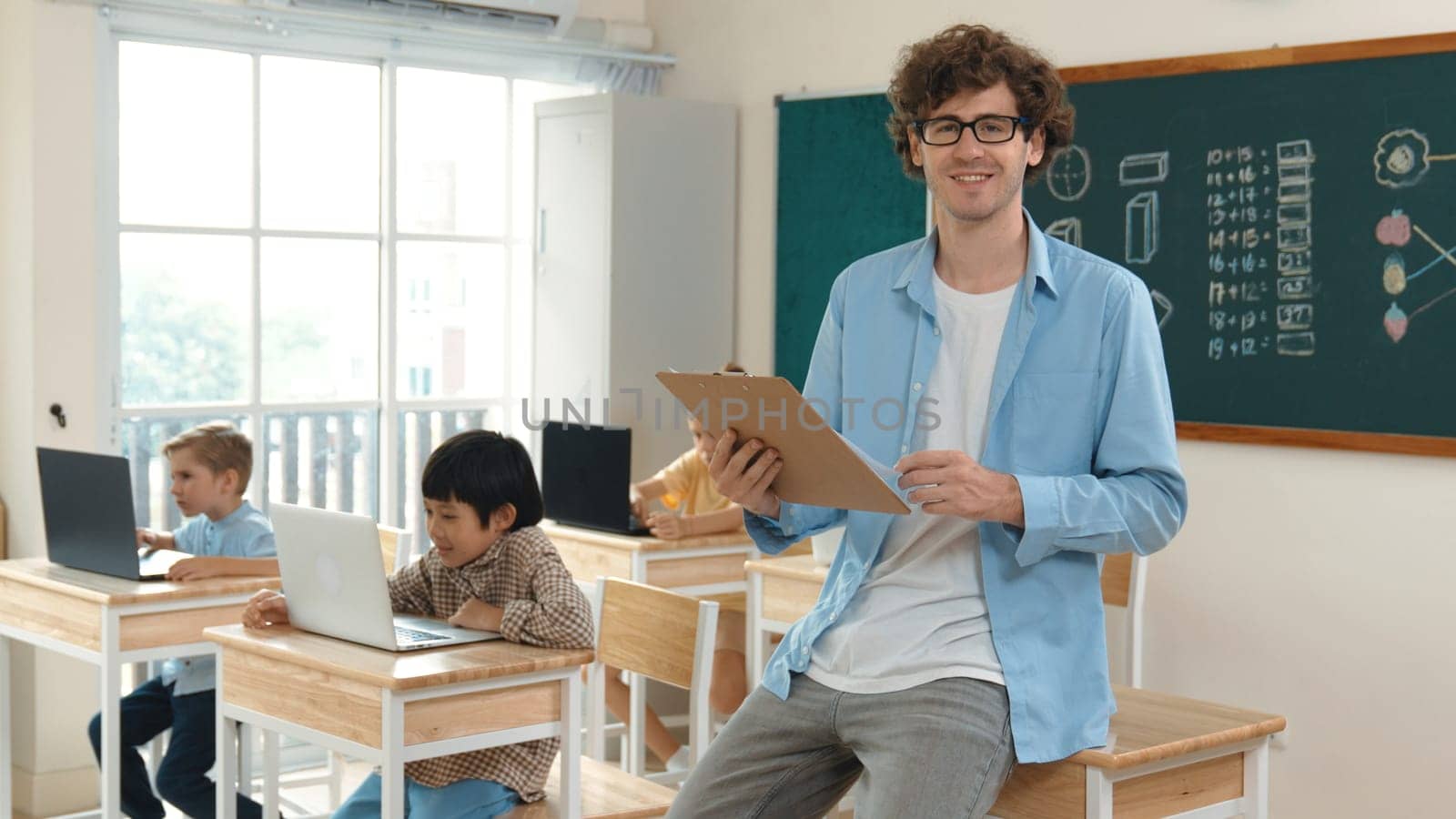 Caucasian teacher checking document and looking at camera at classroom. Diverse children sitting at learning about coding engineering prompt and generated AI. Cute student programing system. Pedagogy.