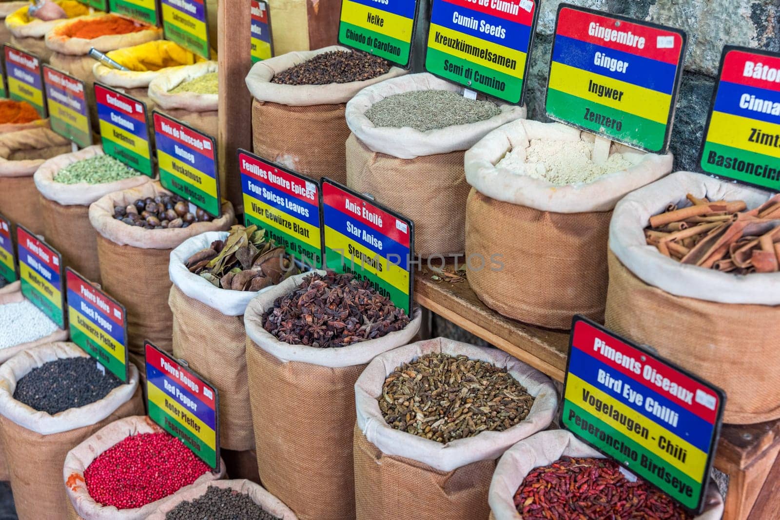 A colorful display of spices and herbs, including cumin, coriander, and ginger. The spices are arranged in small bags and baskets, and there are signs next to each one indicating their names and uses by DaLiu