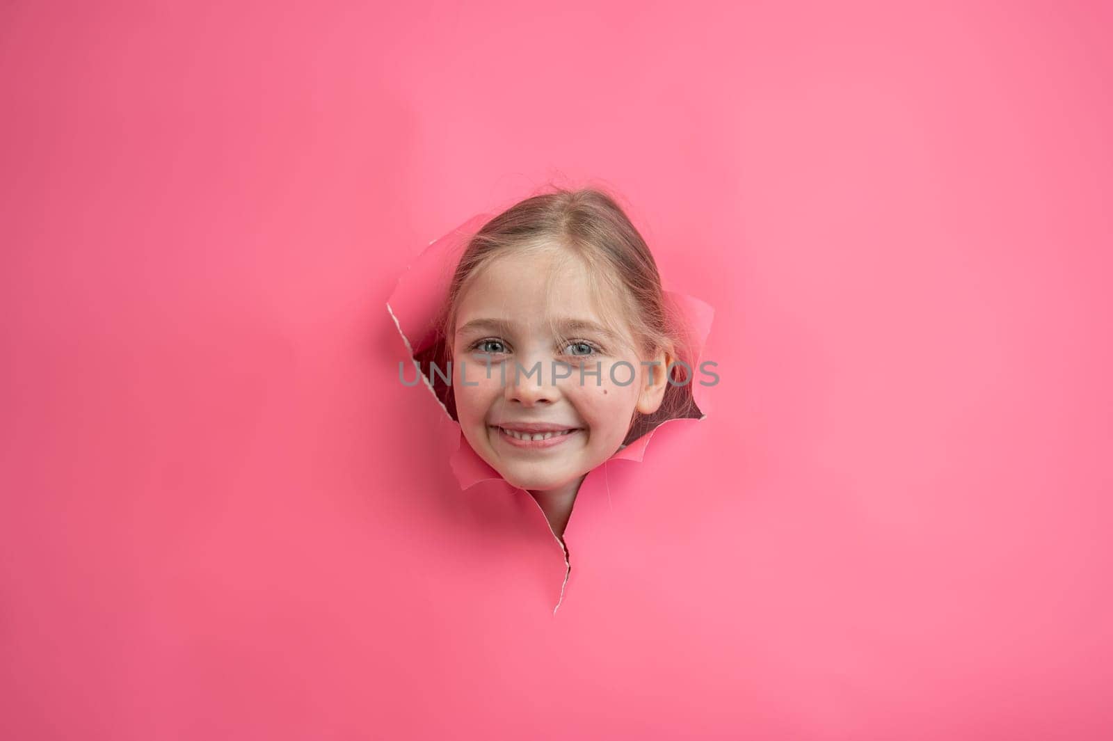 Cute Caucasian girl peeks out of a hole in a paper pink background. by mrwed54