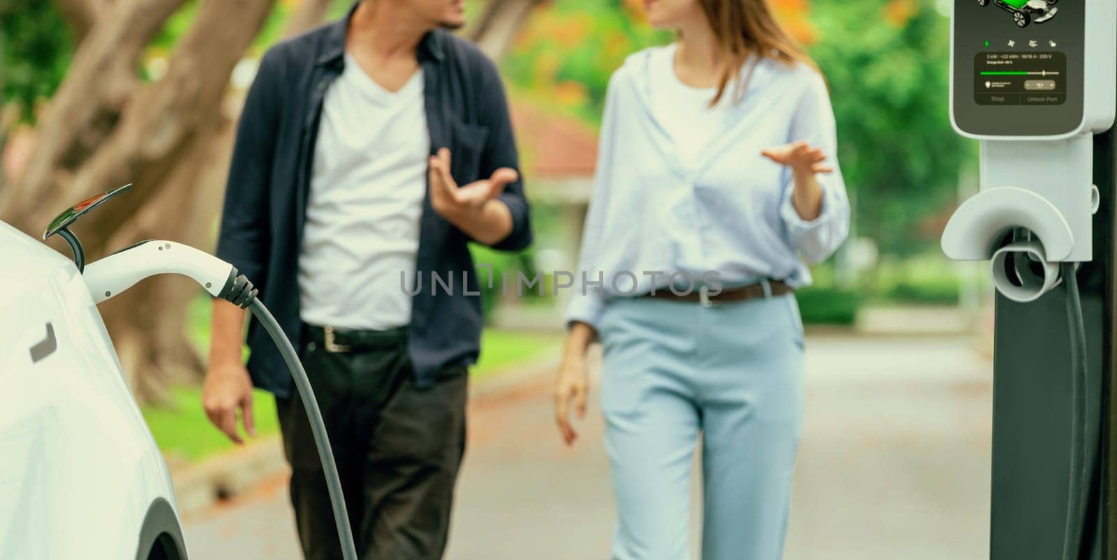 Focused EV car recharging battery on blurred background of lovey couple during autumnal road trip vacation by electric vehicle recharging battery. Eco friendly travel during autumn. Panorama Exalt