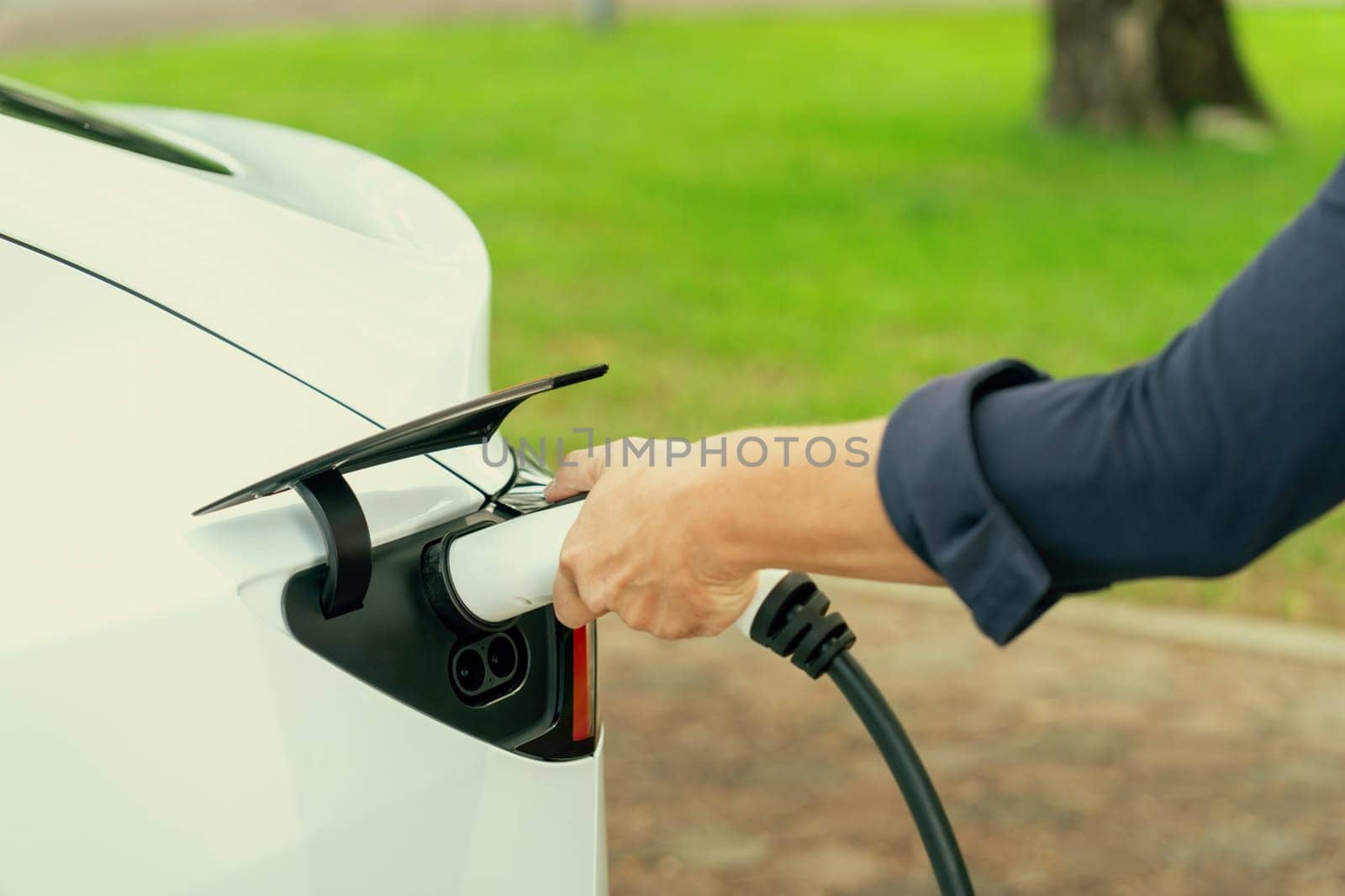 Man recharge EV electric vehicle's battery from EV charging station in outdoor green city park scenic. Eco friendly urban transport and commute with eco friendly EV car travel. Exalt