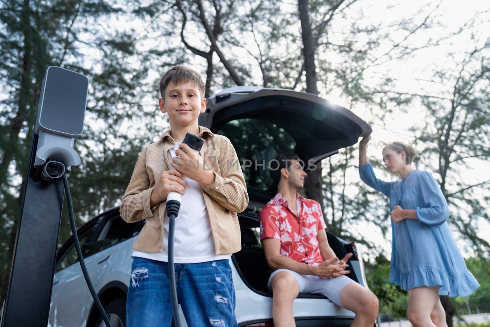 Little boy holding EV charger and point at camera with his family sitting on the trunk in background. Road trip travel with alternative energy charging station for eco-friendly car concept. Perpetual