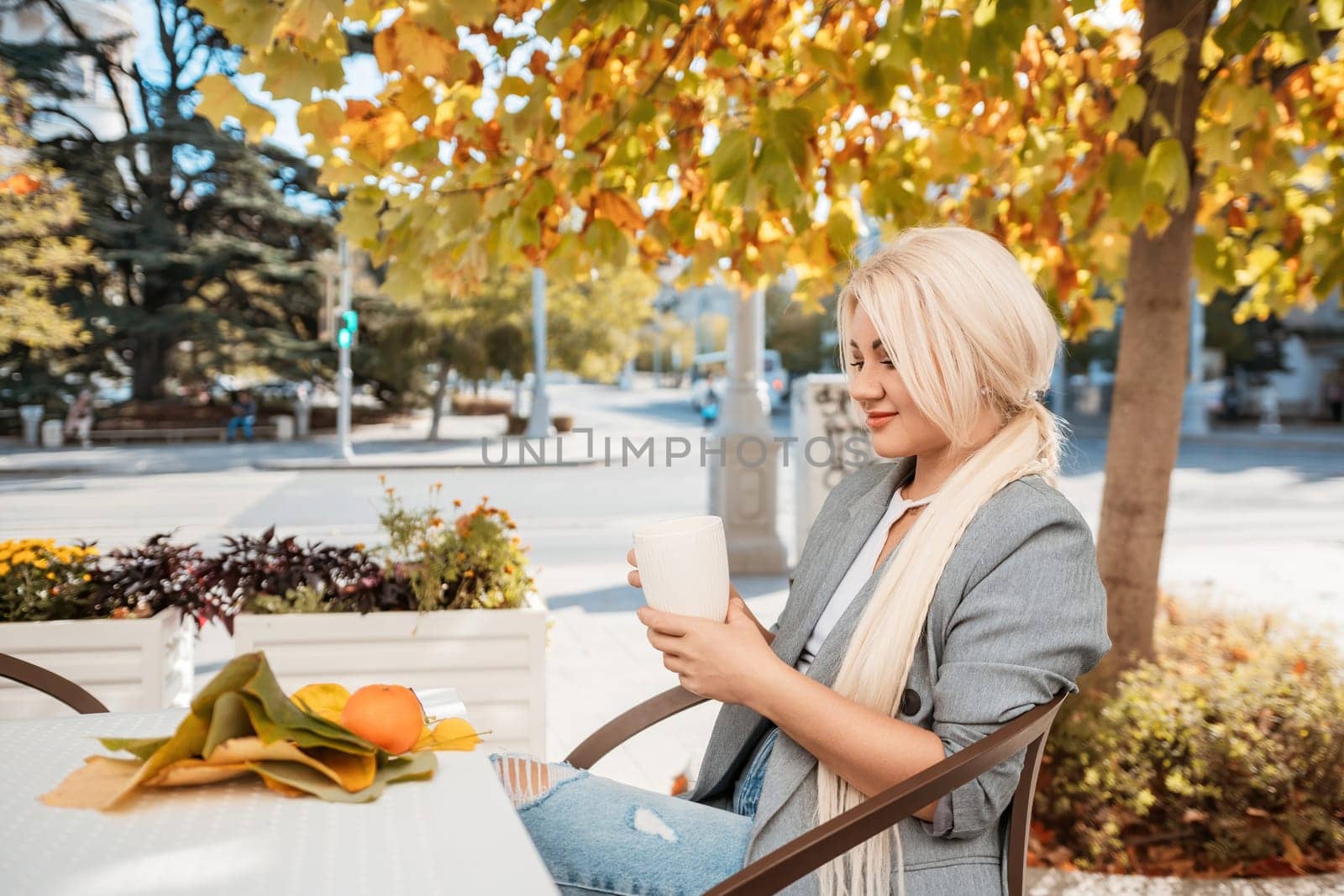 A blonde woman is sitting at a table with a cup of coffee and a plate of fruit. She is smiling and enjoying her time outside