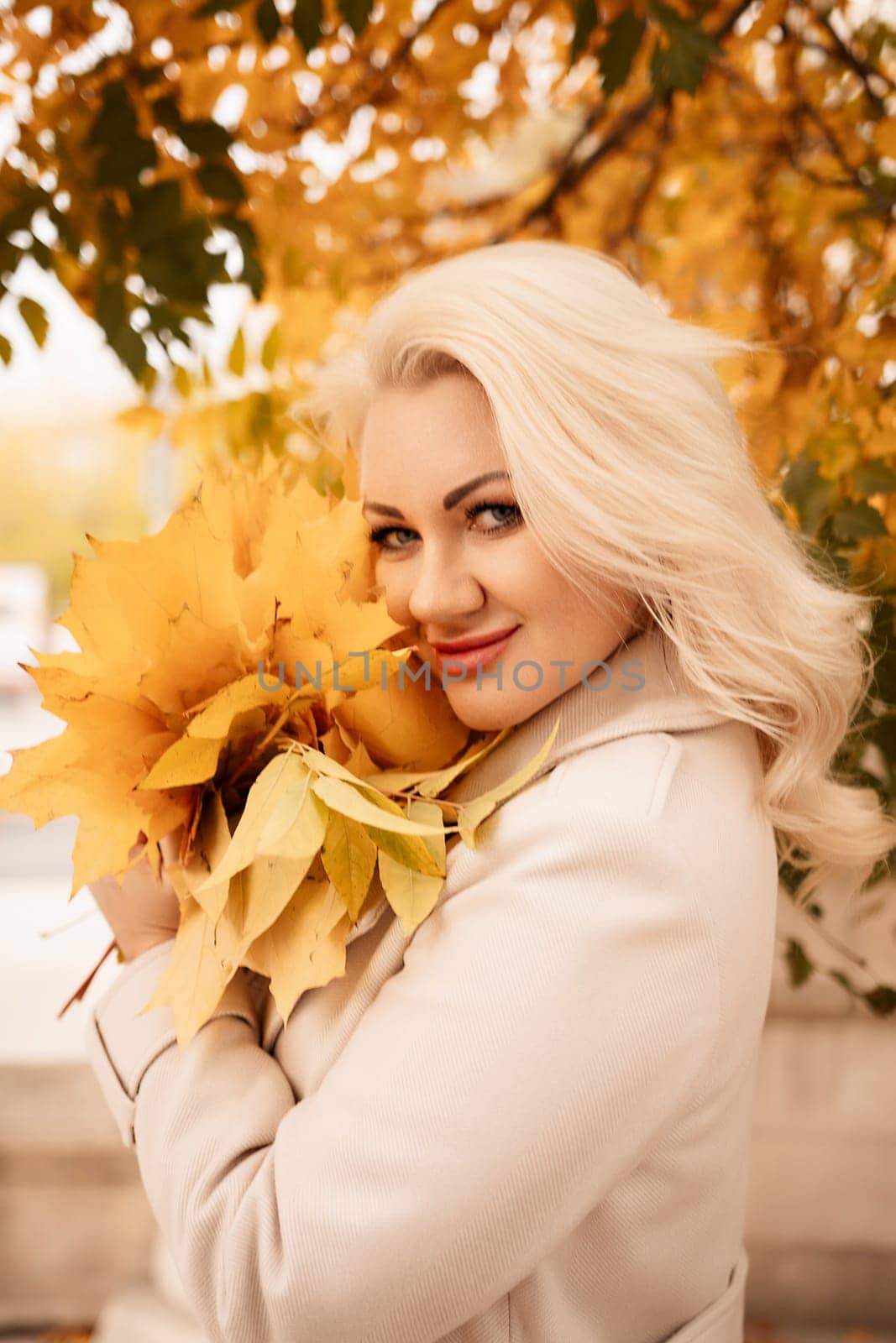 A blonde woman is holding a bouquet of yellow leaves. She is smiling and she is happy. by Matiunina