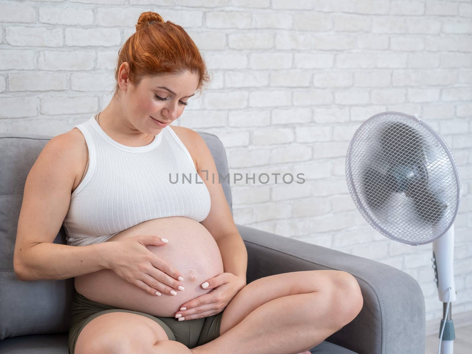 A pregnant woman is sitting on the couch, stroking her tummy and enjoying the cool air from an electric fan. by mrwed54