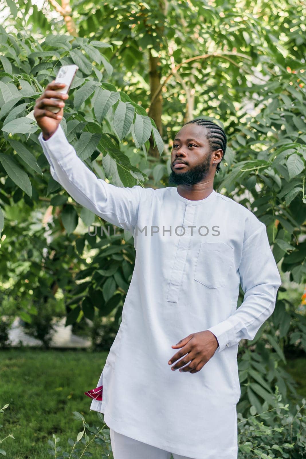 Happy young African American man in dashiki ethnic clothes taking selfie on green background outdoor in summer time. Millennial generation student and youth. by Satura86