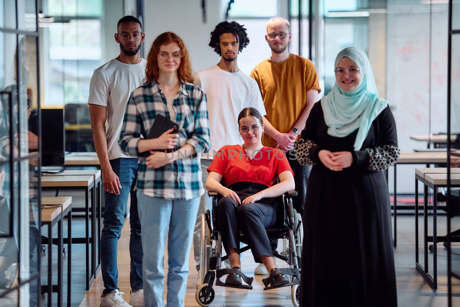 A diverse group of young business people walking a corridor in the glass-enclosed office of a modern startup, including a person in a wheelchair and a woman wearing a hijab.