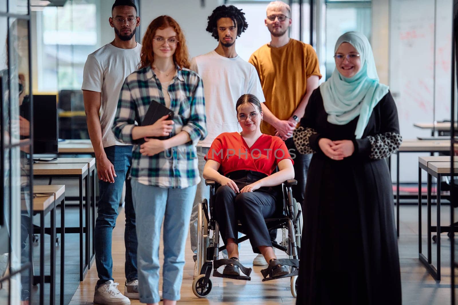 A diverse group of young business people walking a corridor in the glass-enclosed office of a modern startup, including a person in a wheelchair and a woman wearing a hijab.