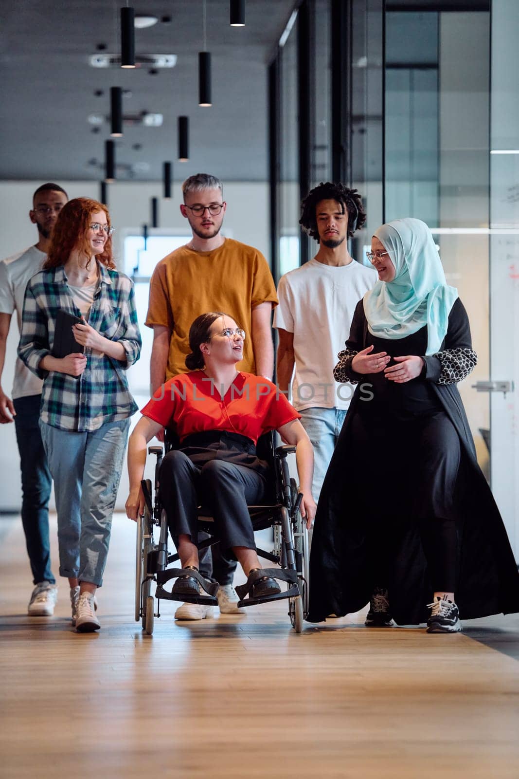 people walking a corridor in the glass-enclosed office of a modern startup, including a person in a wheelchair and a woman wearing a hijab.