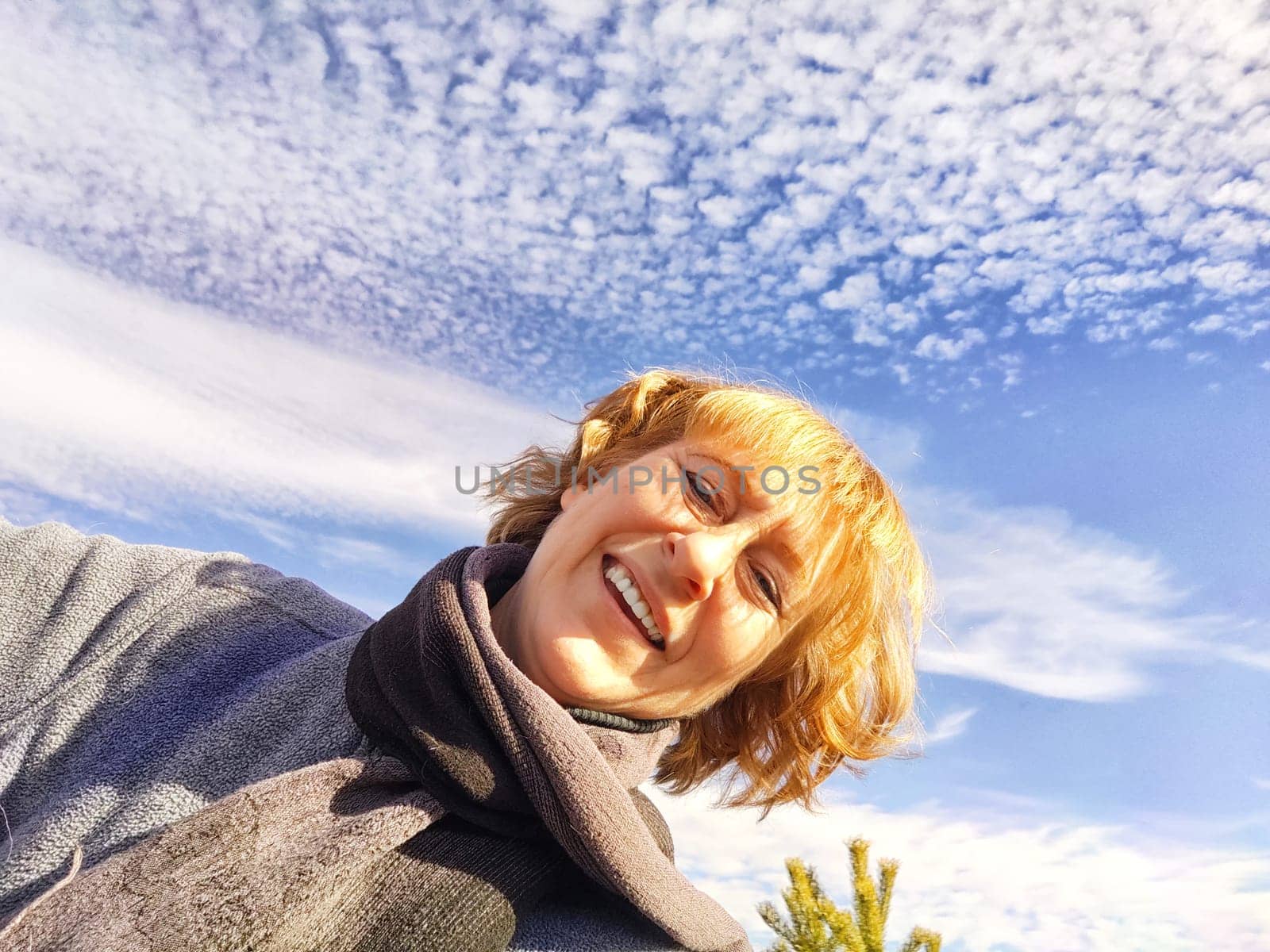 Happy female blonde tourist taking selfie picture with smart mobile phone outside. Middle aged woman having fun on adventure trip and blue sky on background