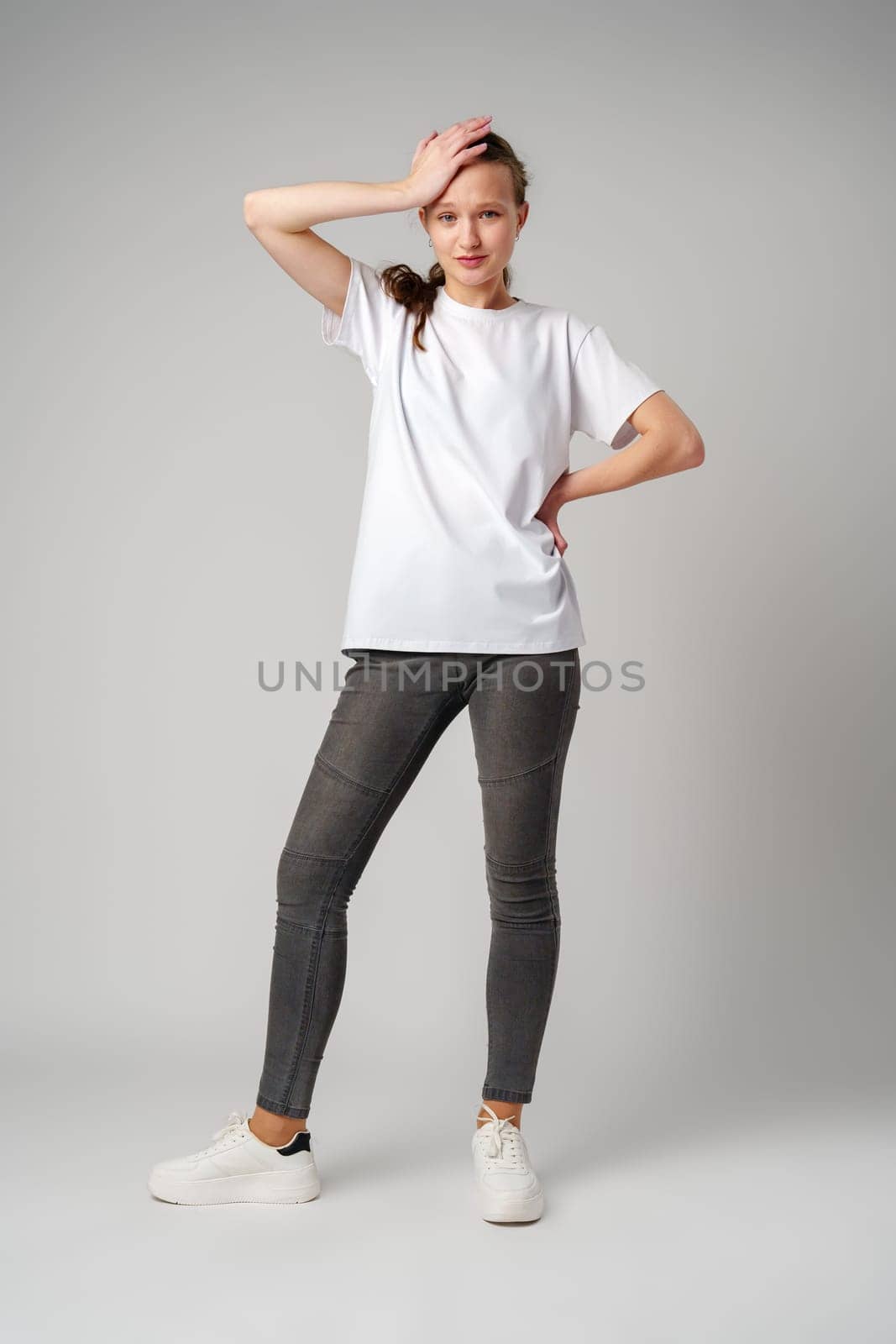 Beautiful young girl posing in white T-shirt and jeans on gray background by Fabrikasimf