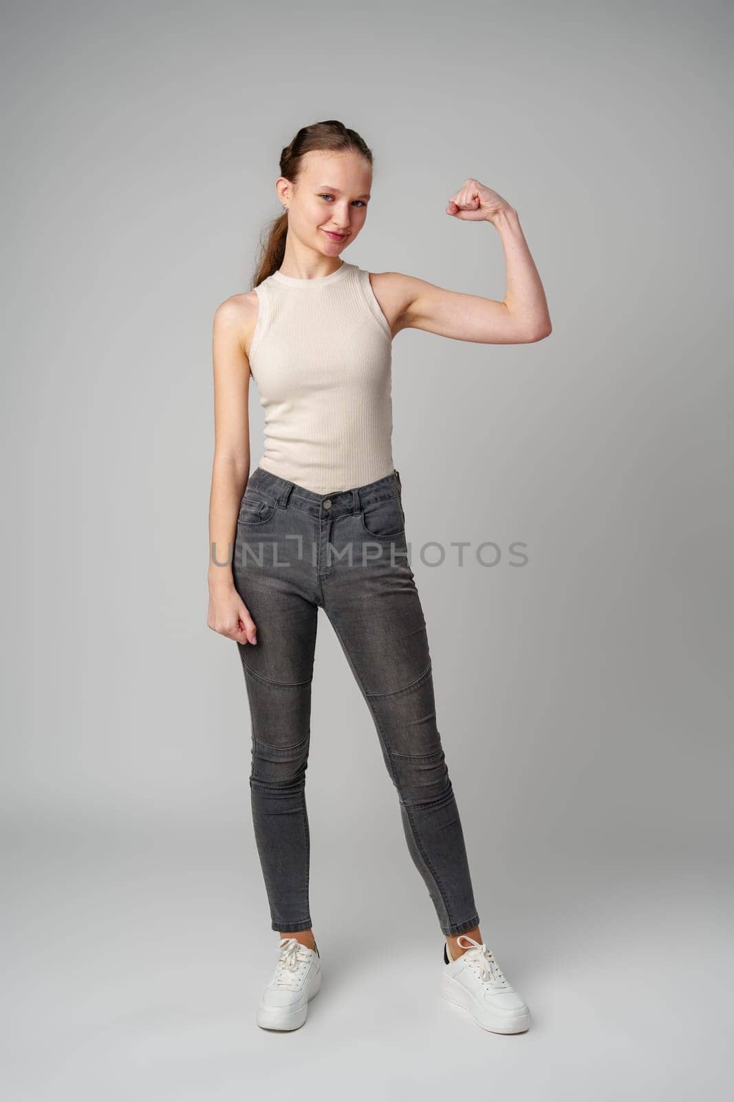 Young Woman Flexing Muscles in Beige Tank Top on gray background in studio