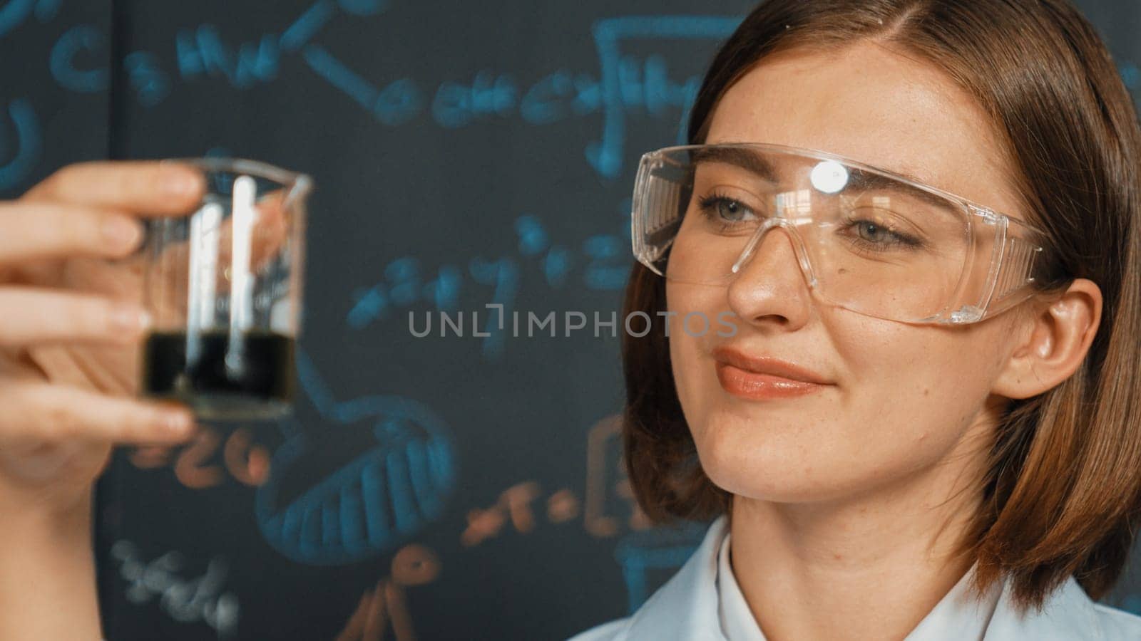 Closeup of young beautiful teacher focus on looking chemical liquid at laboratory. Skilled scientist doing an experiment by inspect colored solution while standing in front of blackboard. Erudition.