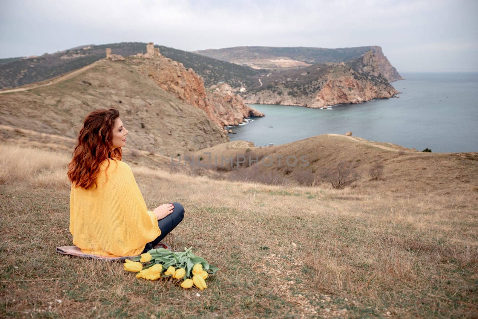 A woman sits on the grass with a bouquet of yellow flowers in her lap. She is looking out at the ocean, taking in the view. Concept of peace and tranquility. by Matiunina