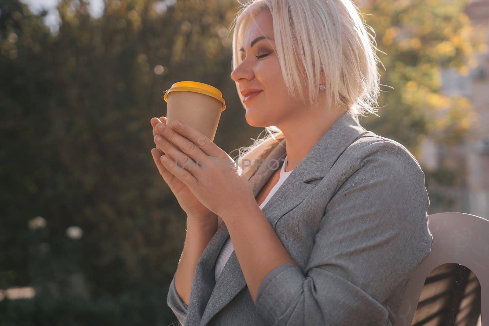 A blonde woman sits on a bench drinking coffee from a yellow cup. She is wearing a gray jacket and has her hair in a ponytail. The scene is peaceful and relaxing