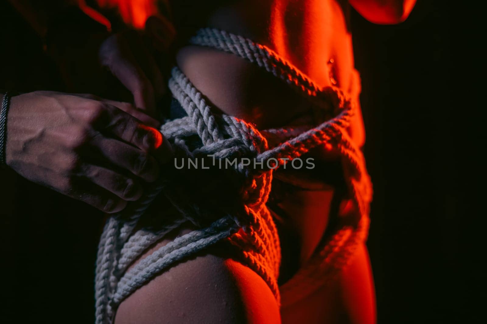 Man tightens ropes using Japanese shibari technique on woman's body in red light