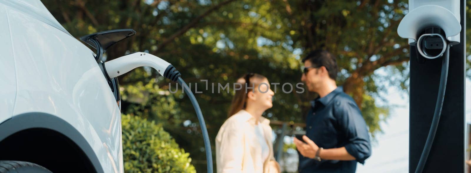 Young couple recharge electric car battery from charging station in green city park in springtime. Rechargeable EV car for sustainable environmental friendly urban travel lifestyle. Panorama Expedient