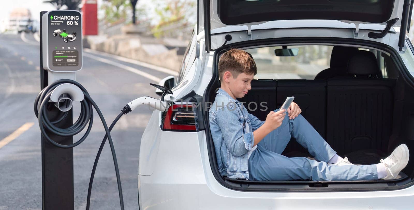 Little boy sitting on car trunk, using smartphone while recharging eco-friendly car from EV charging station. EV car road trip travel as alternative vehicle using sustainable energy concept. Perpetual