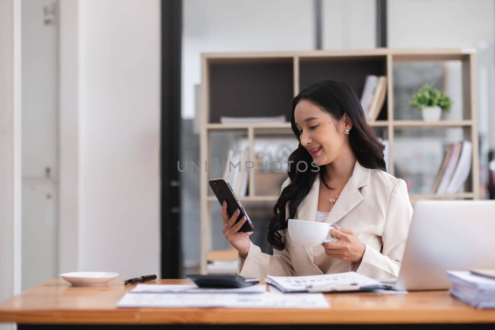 Beautiful business woman explores social media on her phone and drinks coffee before starting work in the morning at her desk. by wichayada