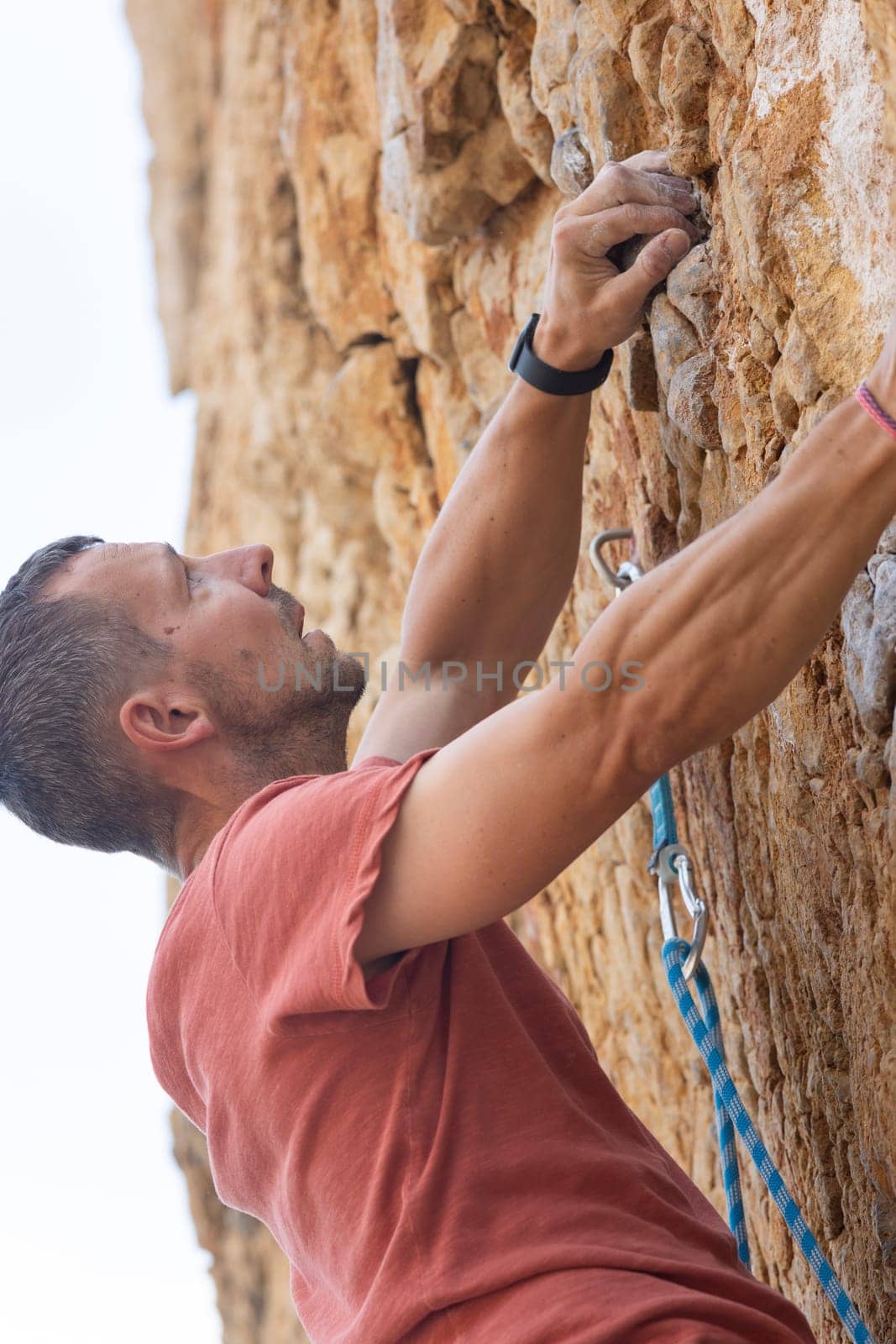 A man in a red shirt is climbing a rock wall. He is wearing a watch and a black wristband