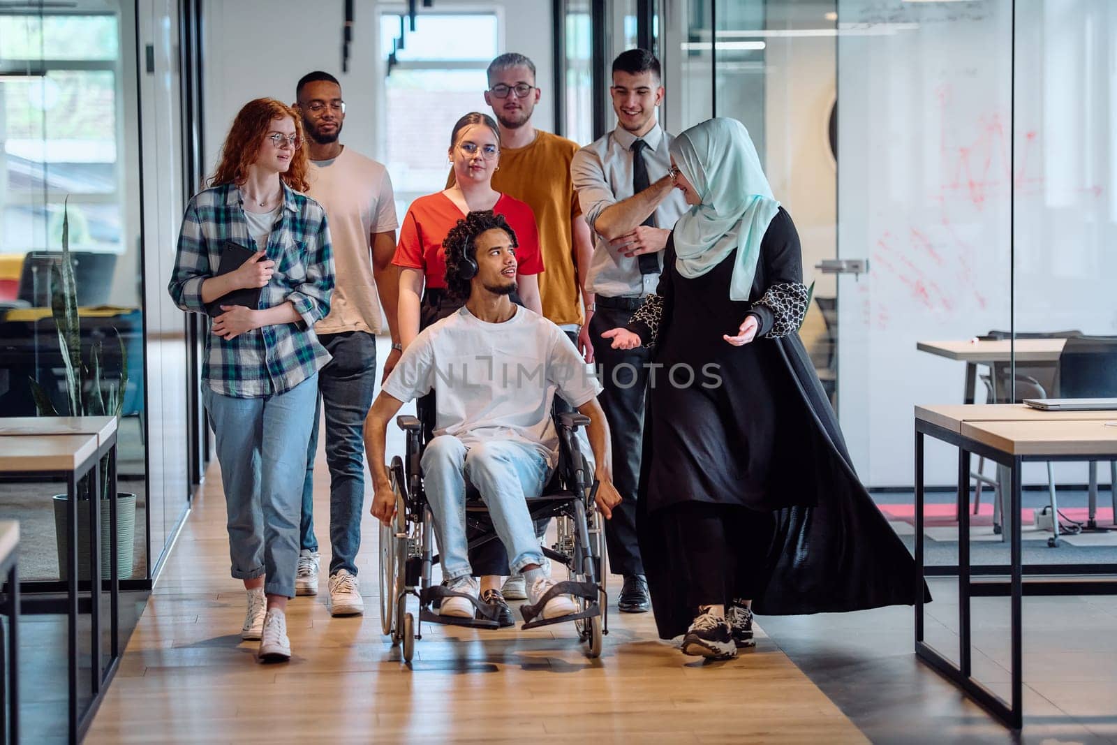 A diverse group of young business people congregates within a modern startup's glass-enclosed office, featuring inclusivity with a person in a wheelchair, an African American young man, and a hijab muslim woman