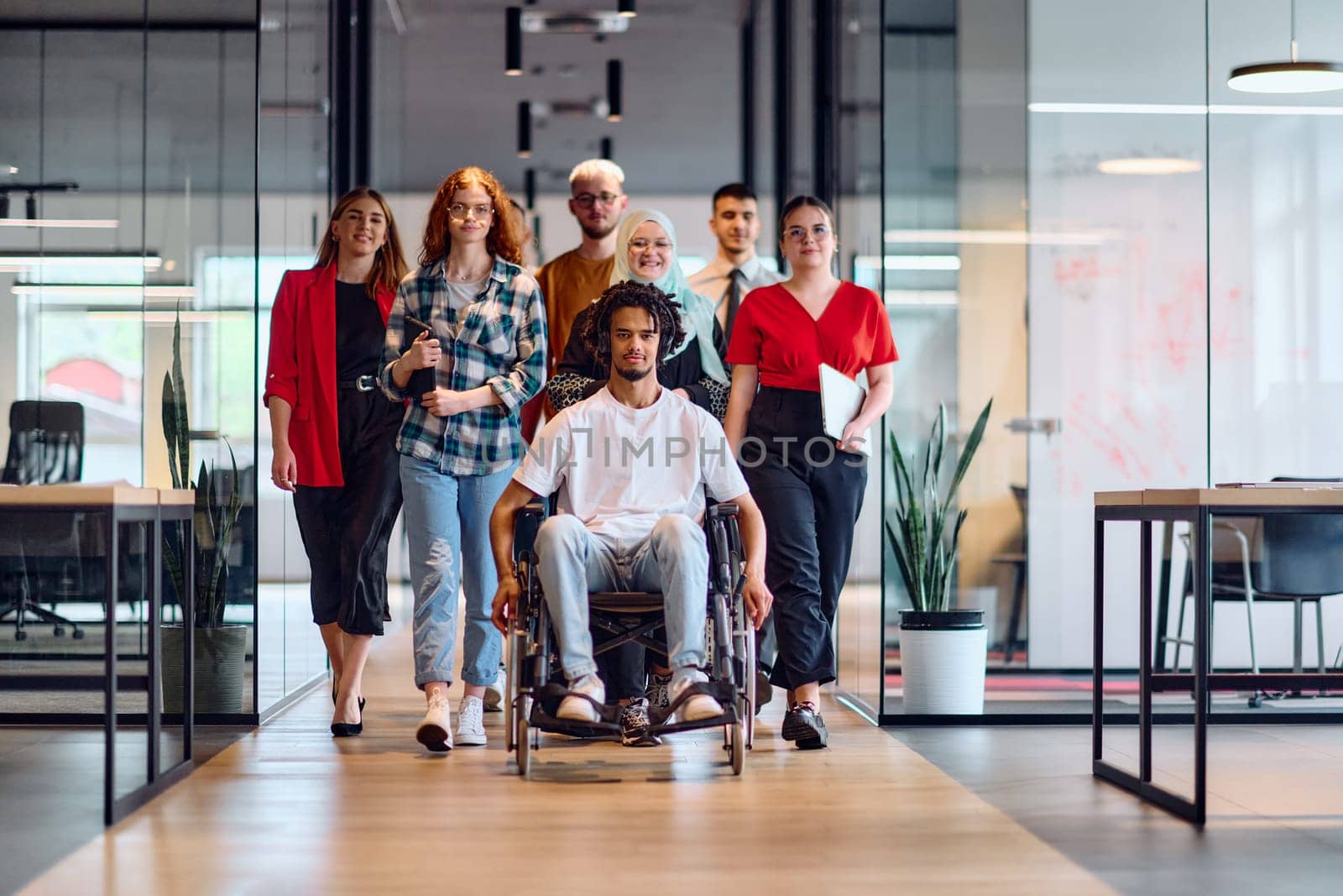 A diverse group of young business people walking a corridor in the glass-enclosed office of a modern startup, including a person in a wheelchair and a woman wearing a hijab.