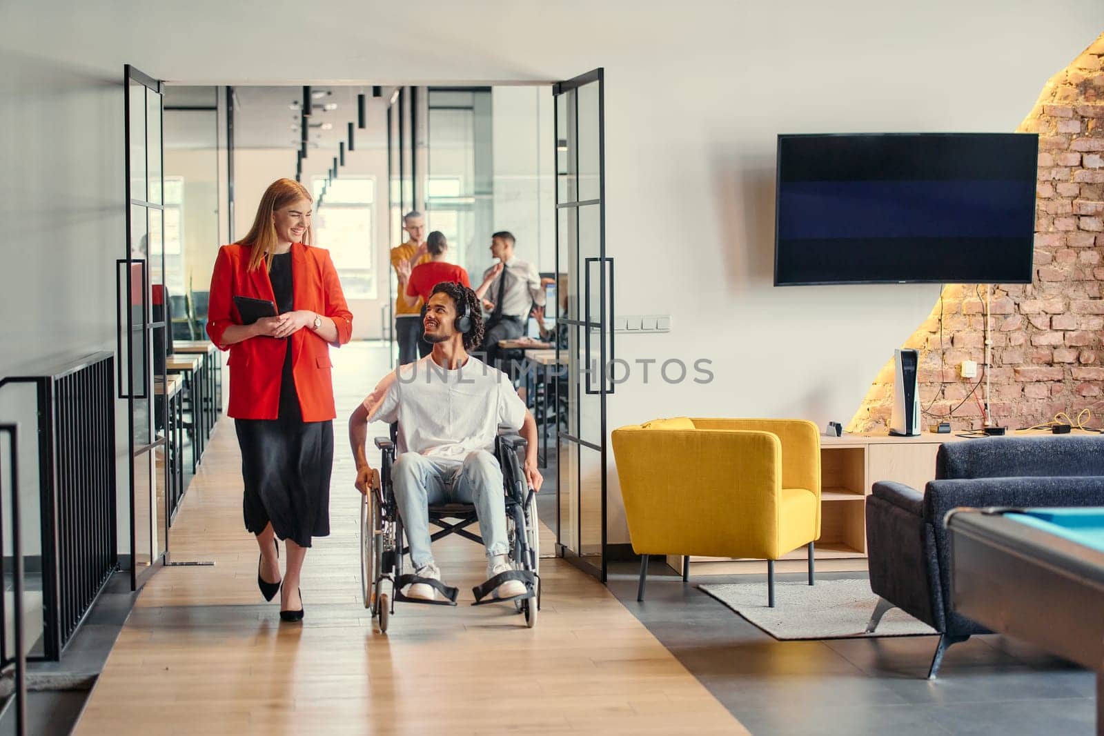 A business leader with her colleague, an African-American businessman who is a disabled person, pass by their colleagues who work in modern offices.