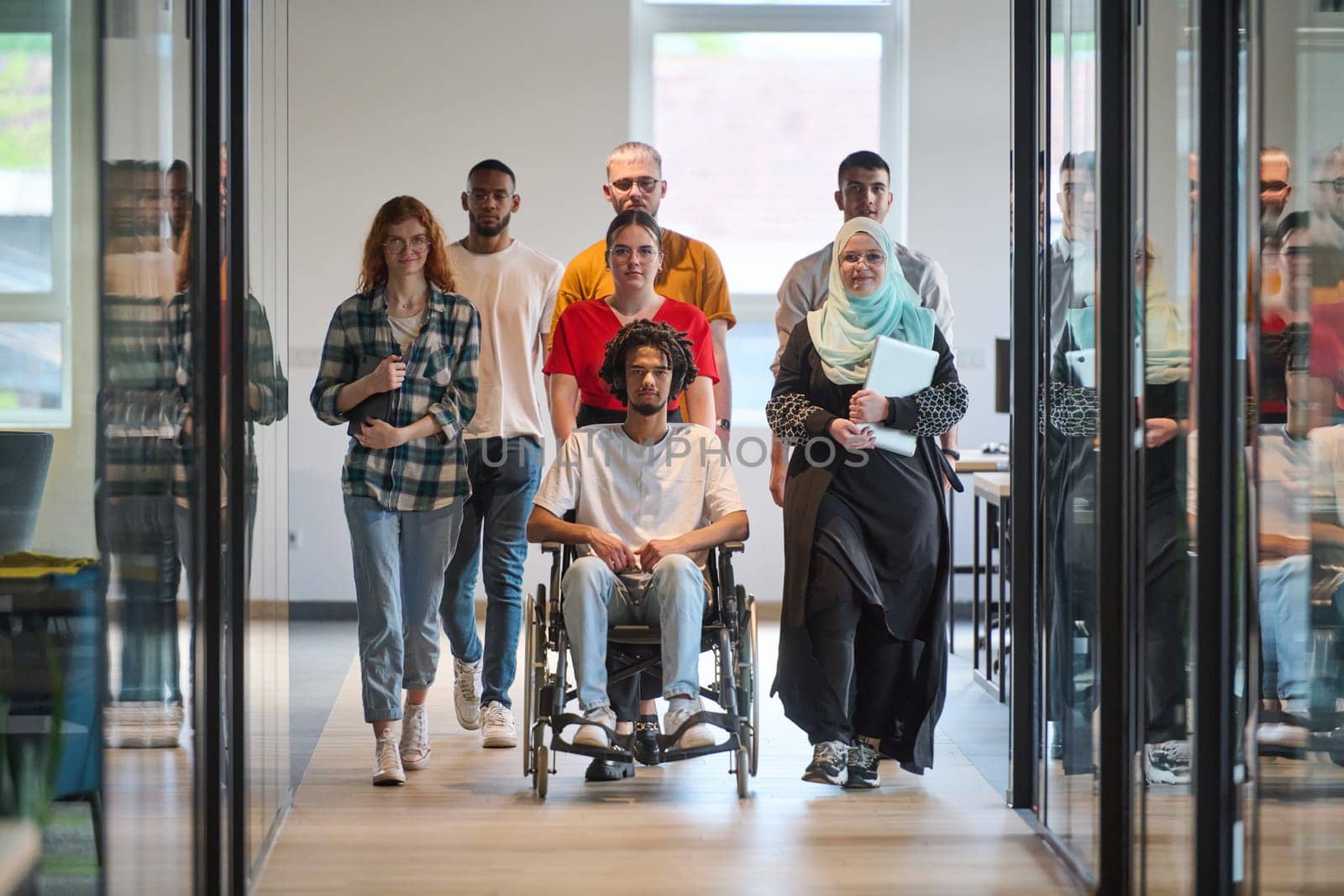 A diverse group of young business people congregates within a modern startup's glass-enclosed office, featuring inclusivity with a person in a wheelchair, an African American young man, and a hijab muslim woman