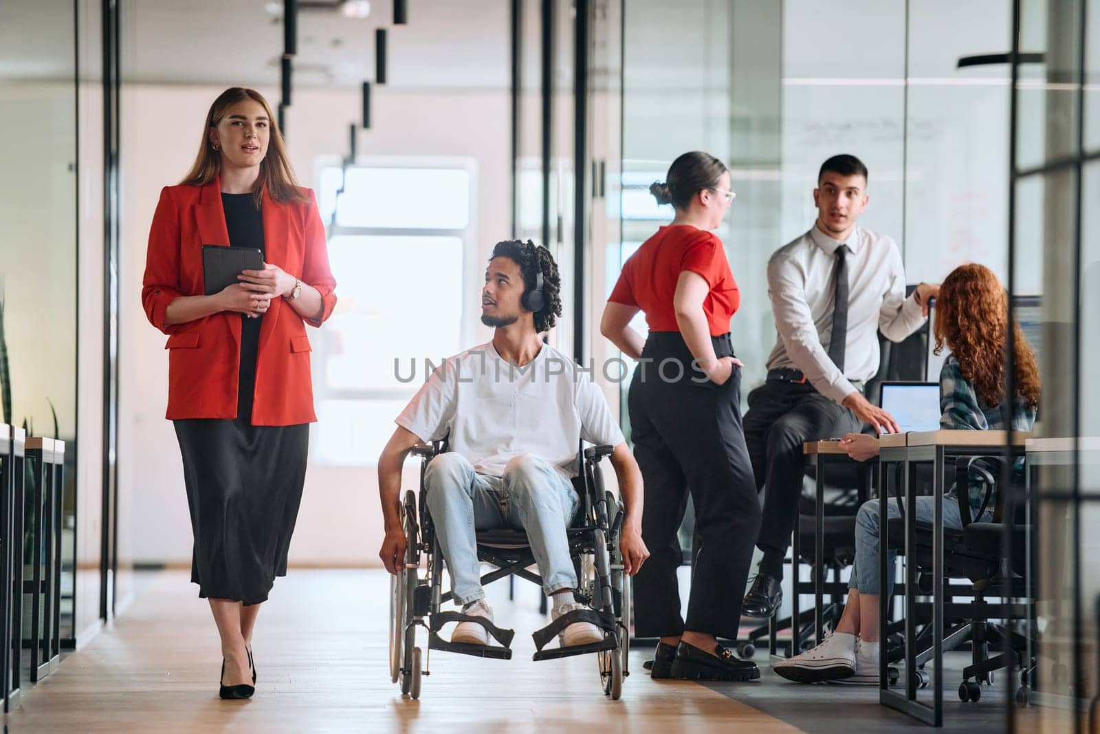 A business leader with her colleague, an African-American businessman who is a disabled person, pass by their colleagues who work in modern offices.