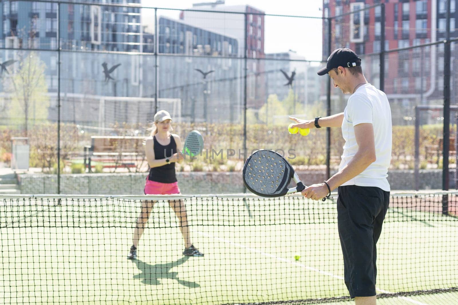 Portrait of positive young woman and adult man standing on padel tennis court, holding racket and ball, smiling by Andelov13