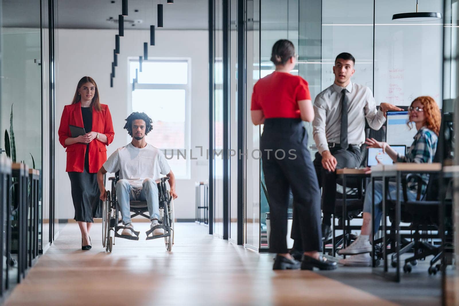 A business leader with her colleague, an African-American businessman who is a disabled person, pass by their colleagues who work in modern offices.